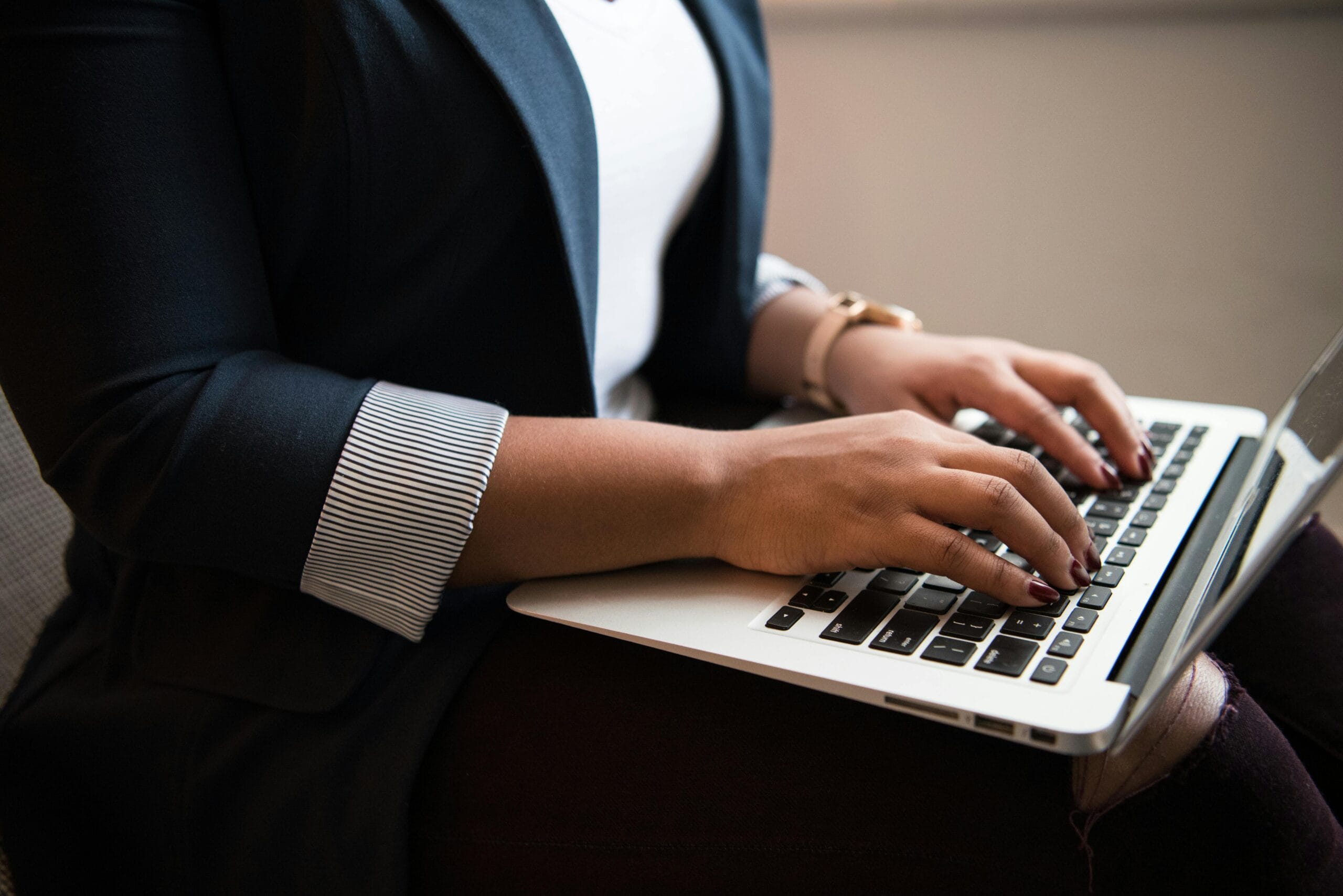Woman in a blazer typing on a laptop in a well-lit room, focused on remote work.